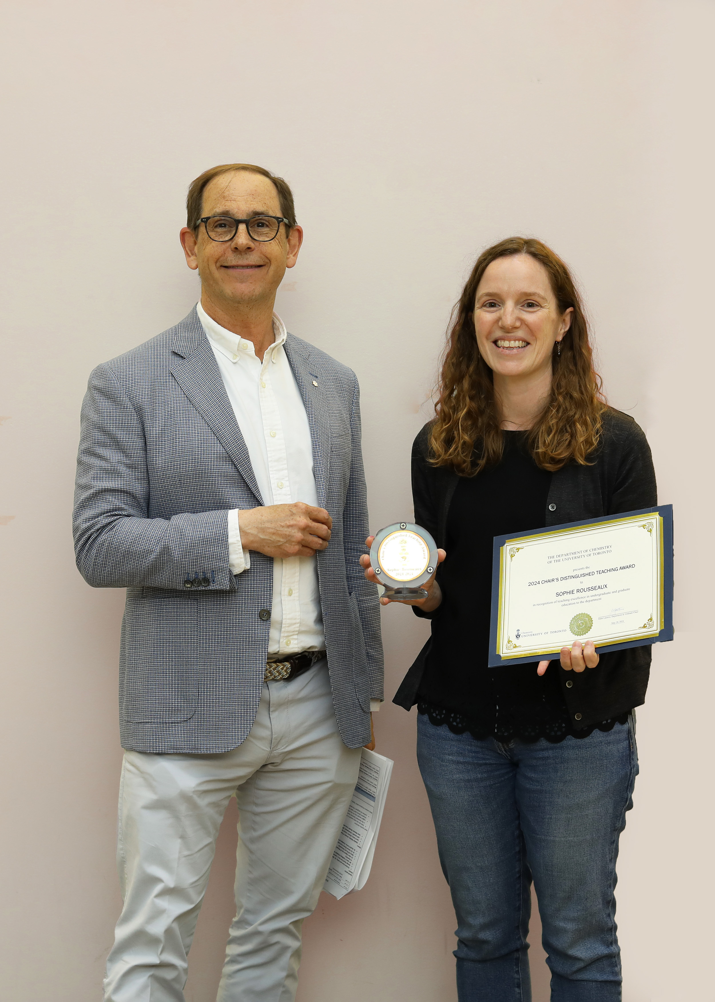 A white man in a suit and a woman with curly red hair stand beside each other, the latter holding a medal and certificate.