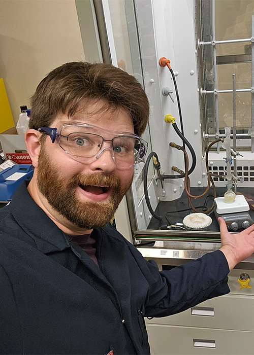 Charlie Kivi, a white man with a beard and moustache, wearing PPE, gestures enthusiastically at lab equipment.