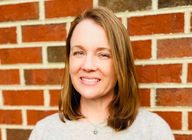 Head shot of a white woman with long, light-brown hair smiling into the camera in front of a brick wall.