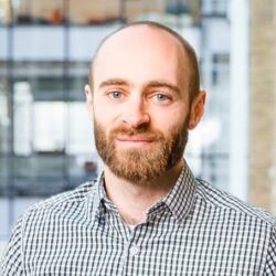 Head shot of Dr. Jon Moir, a white man with a red moustache and beard wearing a mostly white plaid shirt.