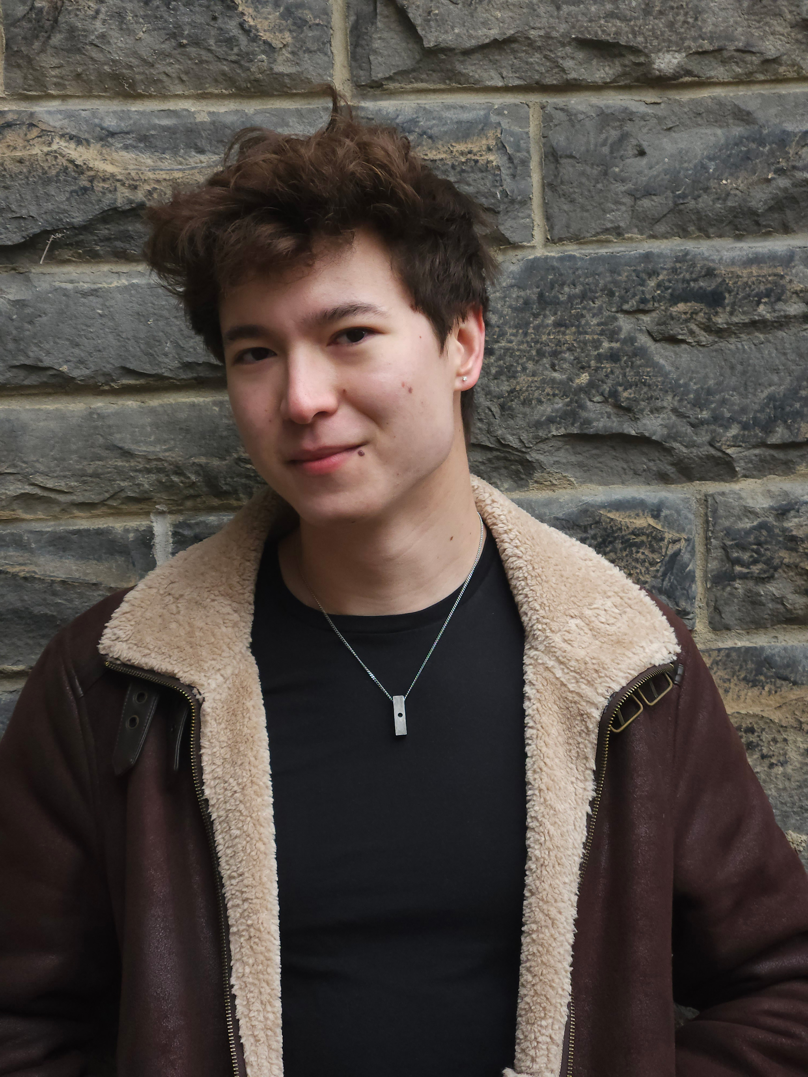 Lenny Chen, a young man with curly brown hair and wearing a bomber jacket, poses in front of a grey stone wall.