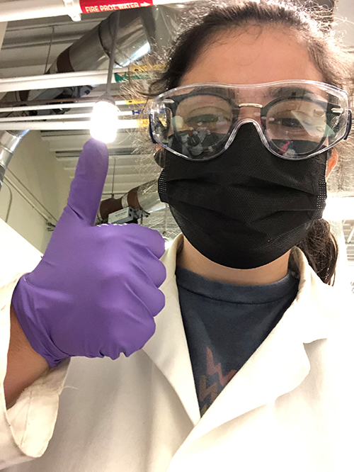 A woman in lab coat, goggles and a mask gives a thumbs up to the camera.