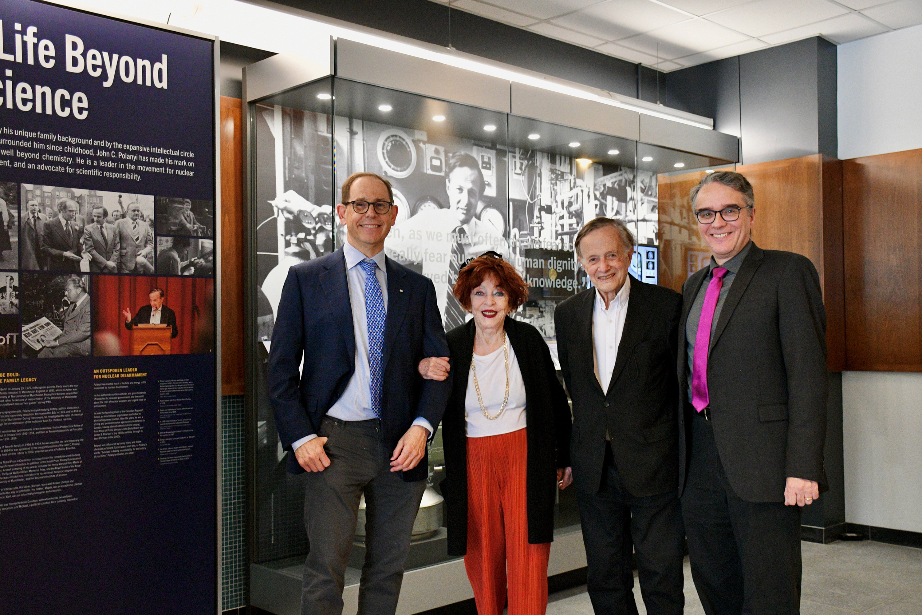 A man, woman and two more men gather with linked arms in front of a museum display, smiling at the camera.