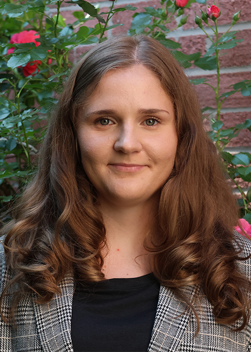 Head shot of chemistry student Yulia Tobolvskaya, a white woman with wavy, shoulder-length brown hair, standing amid roses in front of a brick wall.