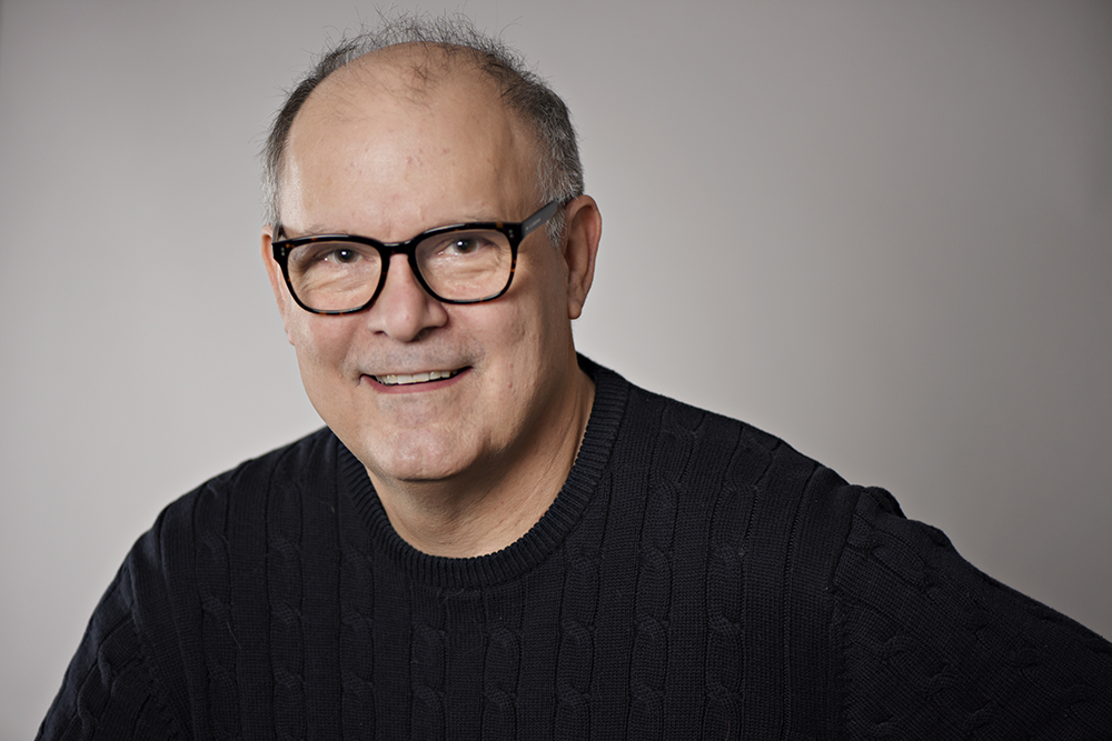 Head shot of a middle aged white man with glasses, wearing a black jumper in front of a grey background.