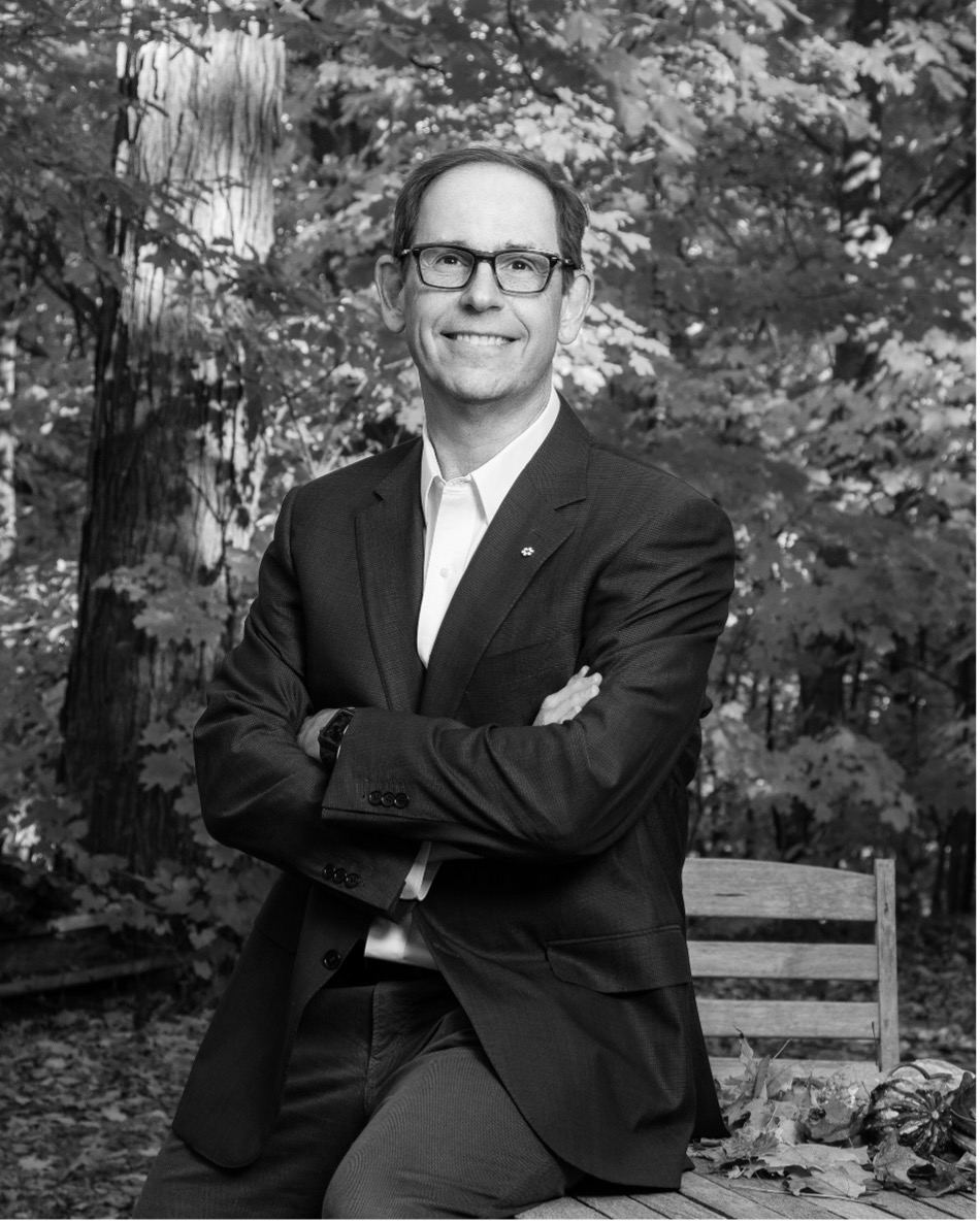 A white man with glasses and a suit poses in front of a wooded area. Picture is black and white.
