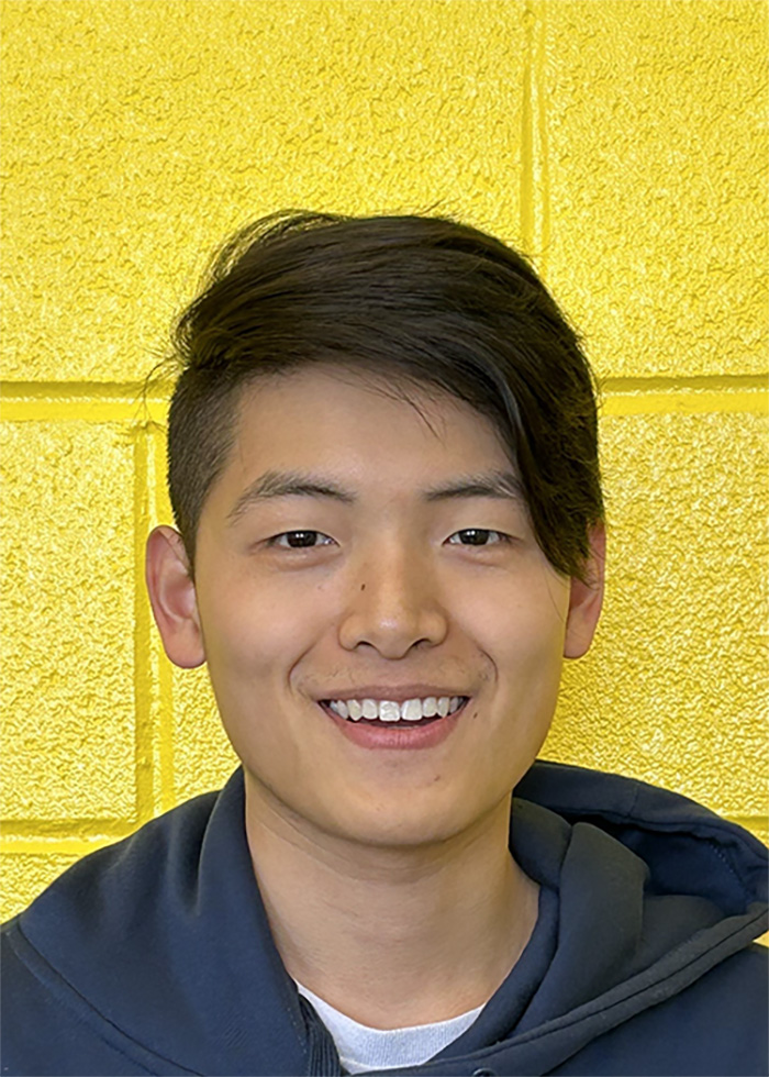 Head shot of a smiling Asian man in his twenties standing in front of a yellow wall.