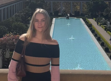 A blonde woman in a striped dress stands in front of a classical garden and long artificial pool.