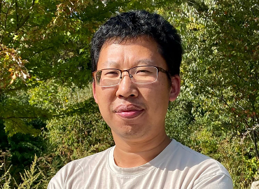 An Asian man with crossed arms stands outside in front of some trees.