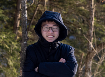 A smiling Asian man with glasses and a hooded winter coat stands, arms crossed, in front of a wooded area.