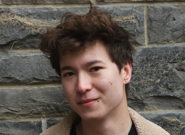 Lenny Chen, a young man with curly brown hair and wearing a bomber jacket, poses in front of a grey stone wall.