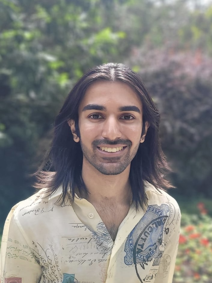 Portrait of a South Asian man with shoulder-length brown hair, smiling at the camera.